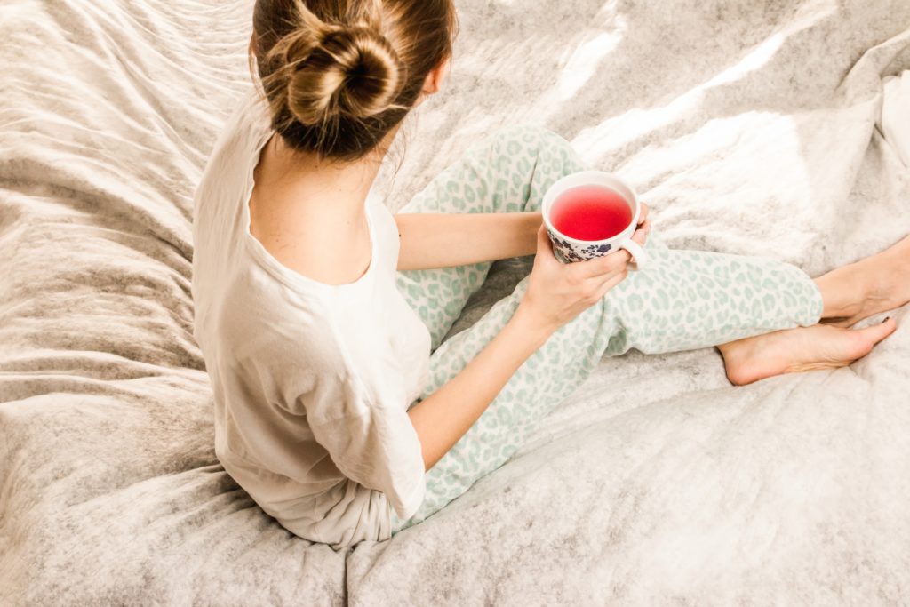 A woman sitting on her bed holding a cup of tea and practicing self-care